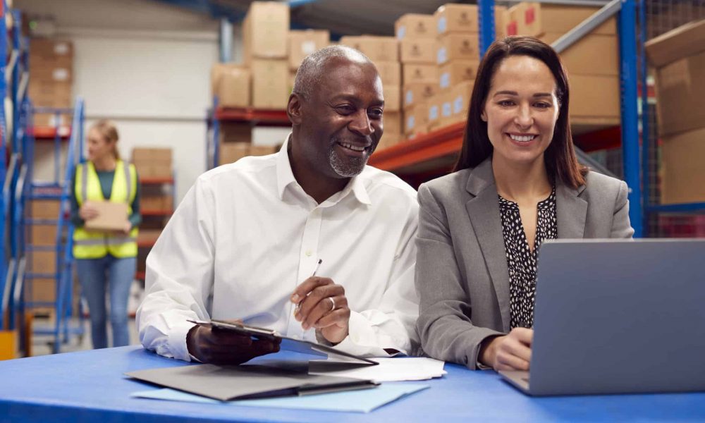 Male And Female Team Leaders Working On Laptop In Warehouse With Workers In Background